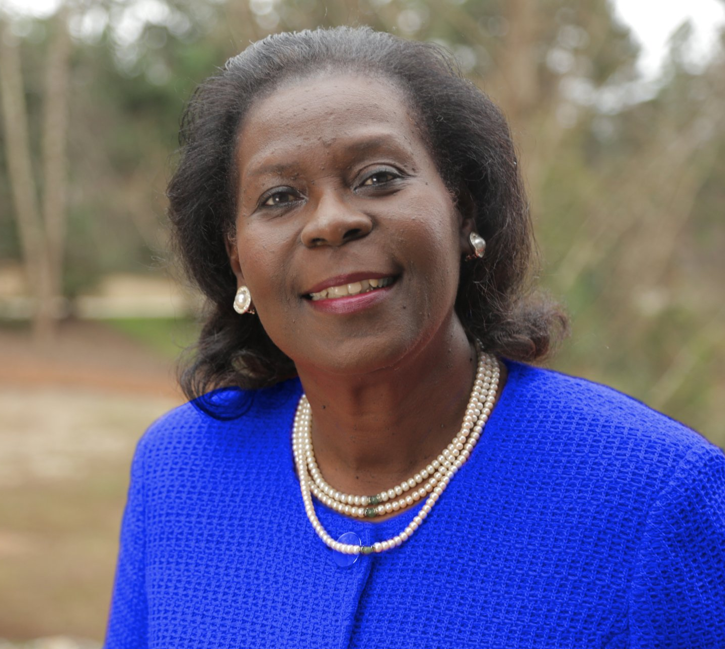 A woman wearing a blue jacket and pearls is smiling for the camera.