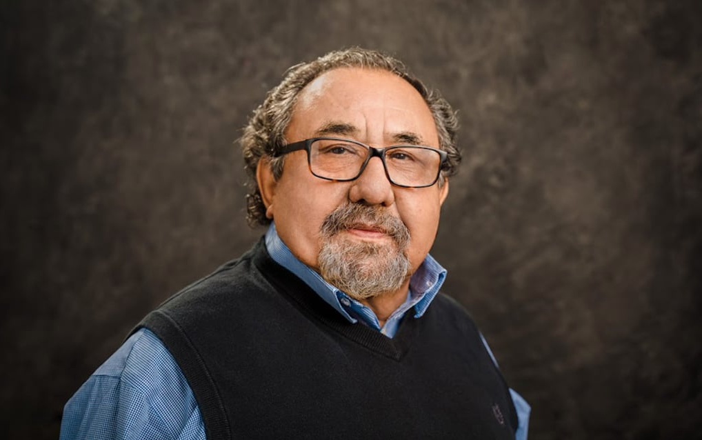 A man with glasses and a beard is wearing a blue shirt and a black vest.