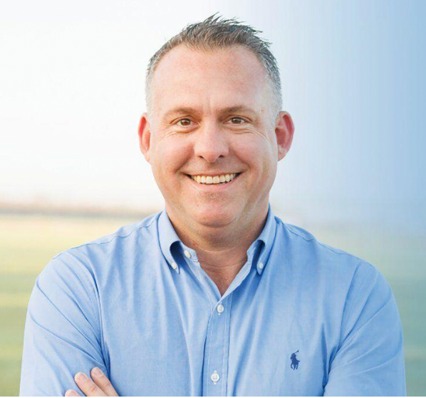 A man in a blue polo shirt is smiling with his arms crossed