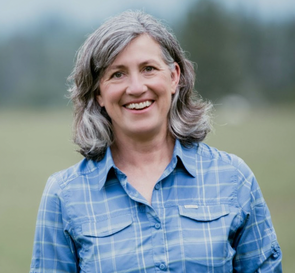 A woman with gray hair is wearing a blue plaid shirt and smiling.