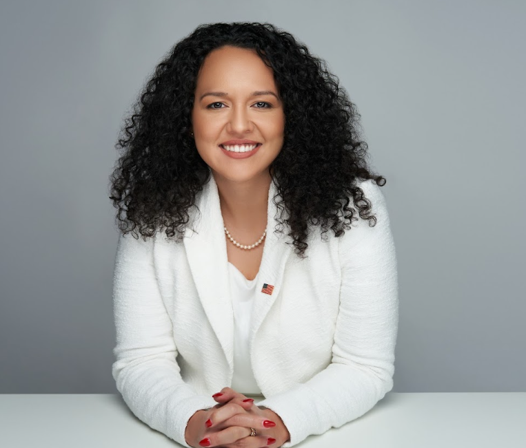 A woman with curly hair is sitting at a table with her hands folded and smiling.