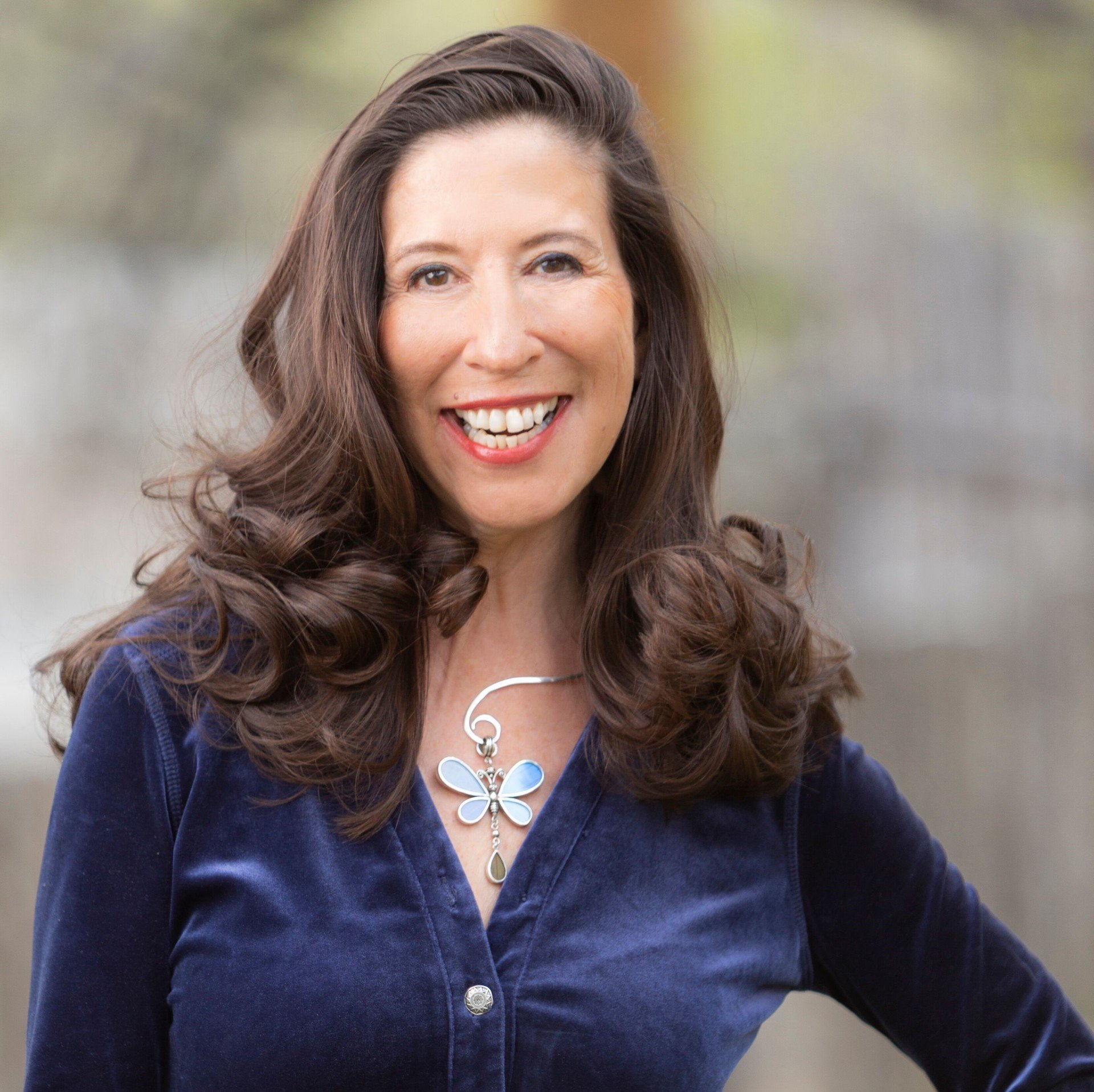 A woman wearing a blue cardigan and a dragonfly necklace smiles for the camera.