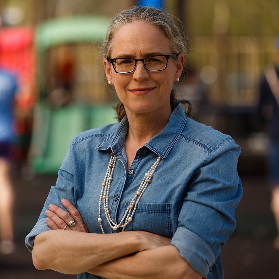 A woman wearing glasses and a denim shirt is standing with her arms crossed.