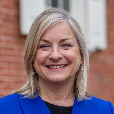 A woman in a blue jacket is smiling in front of a brick building.