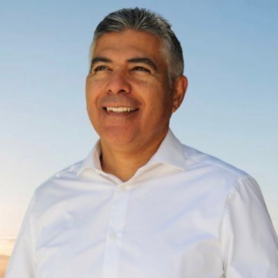 A man in a white shirt is smiling in front of a blue sky.