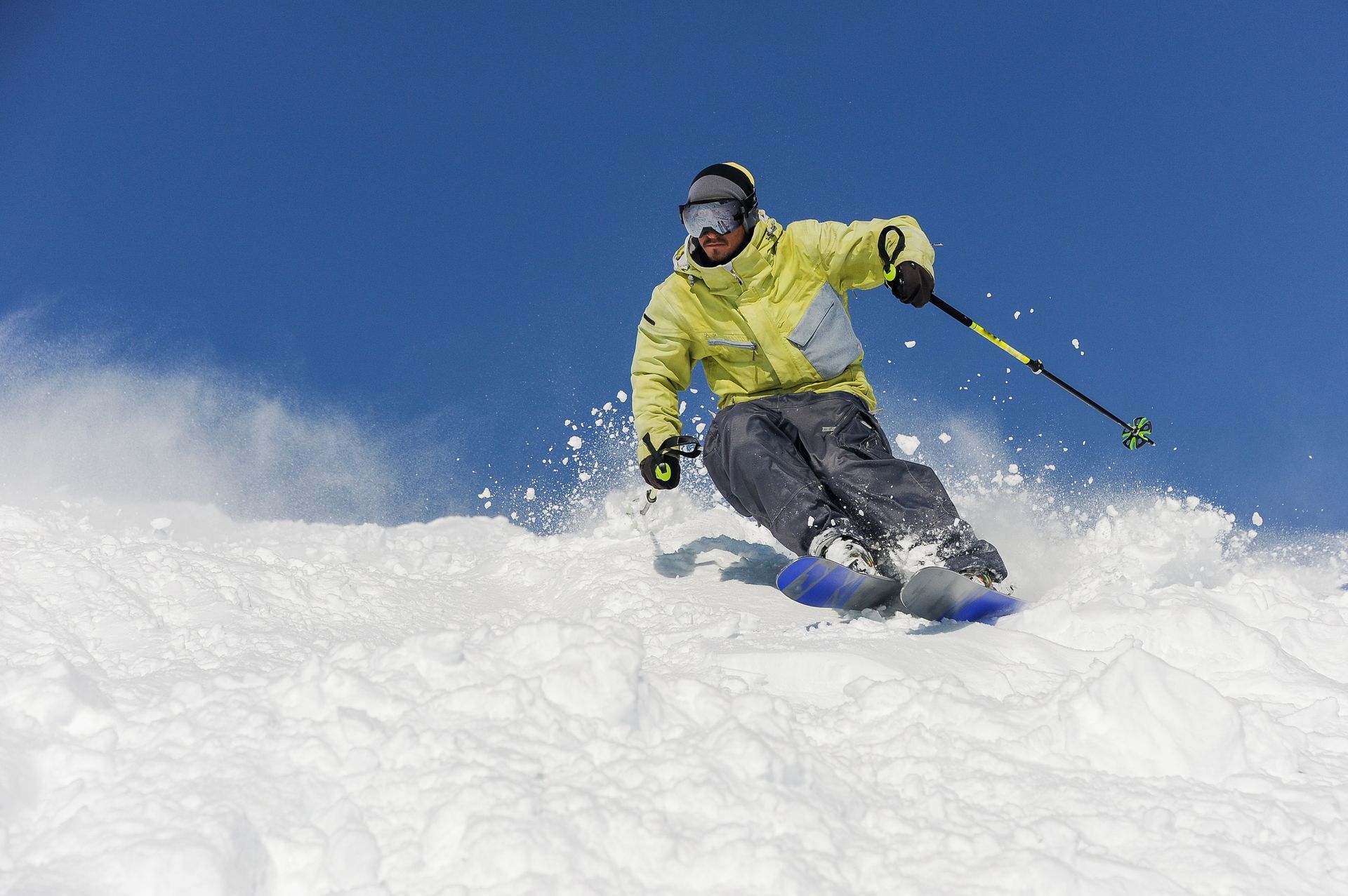 Skier in yellow jacket carving down a snowy slope against a blue sky, creating a spray of snow.