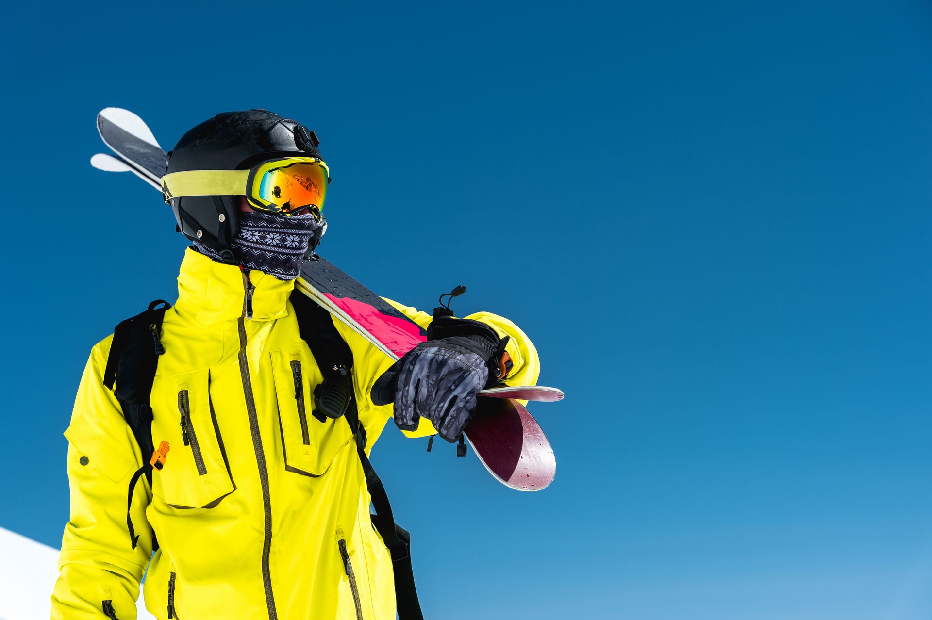 Skier in yellow jacket, black helmet, and goggles, holding skis against a blue sky.