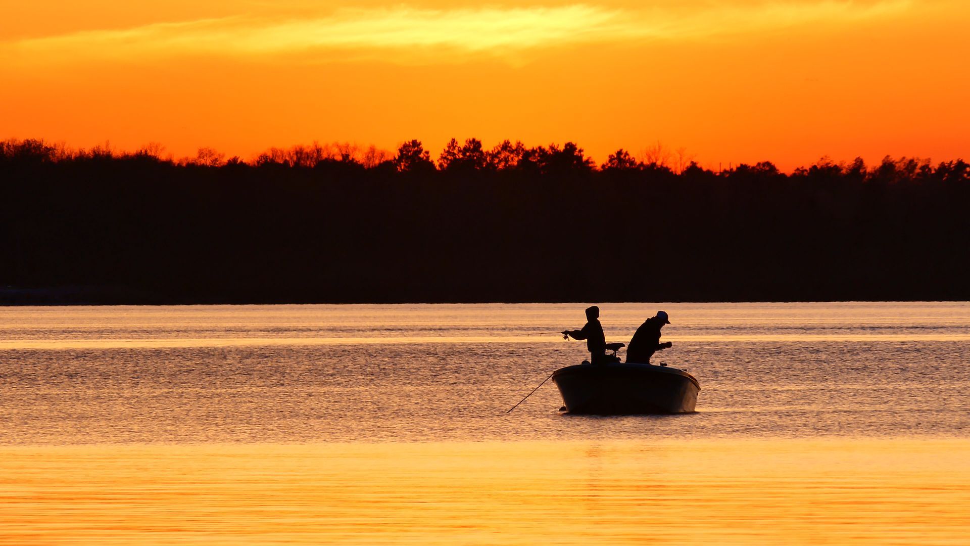 Silhouetted boat with two people fishing on a lake at sunset; orange sky and trees in the background.