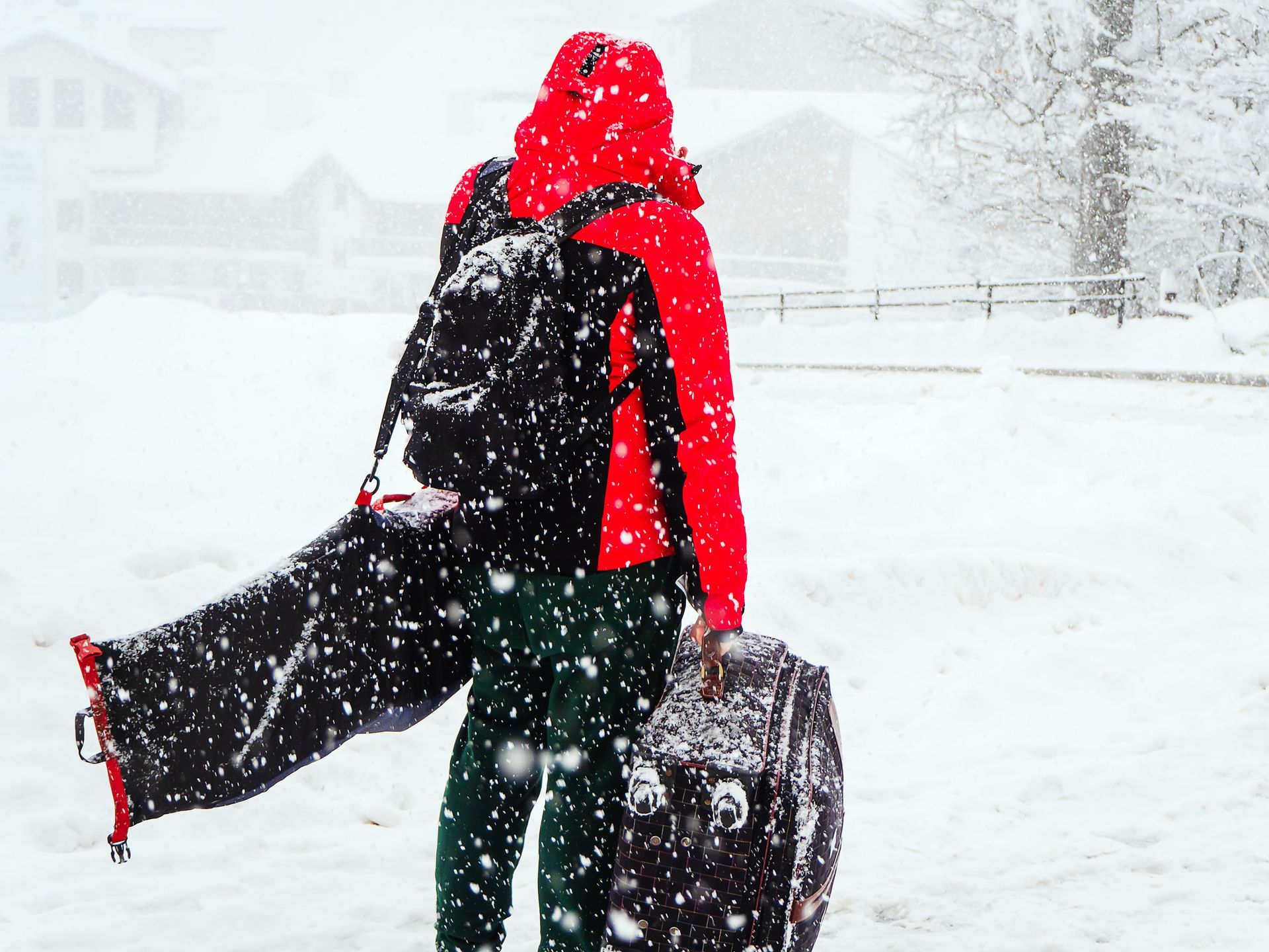 Person bundled in red jacket, backpack, and luggage walking in heavy snow.