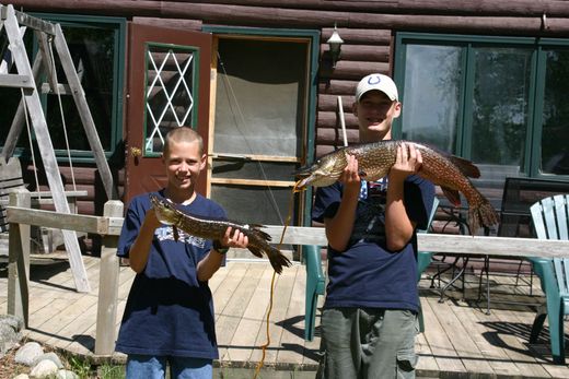 Two boys holding a large fish in front of a log cabin