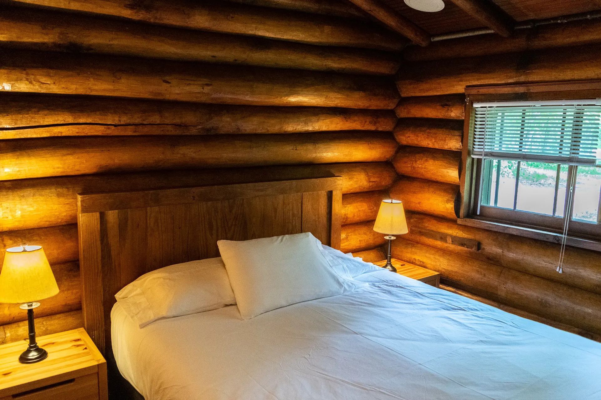 Bedroom in a log cabin with a bed, nightstands, lamps, and a window.