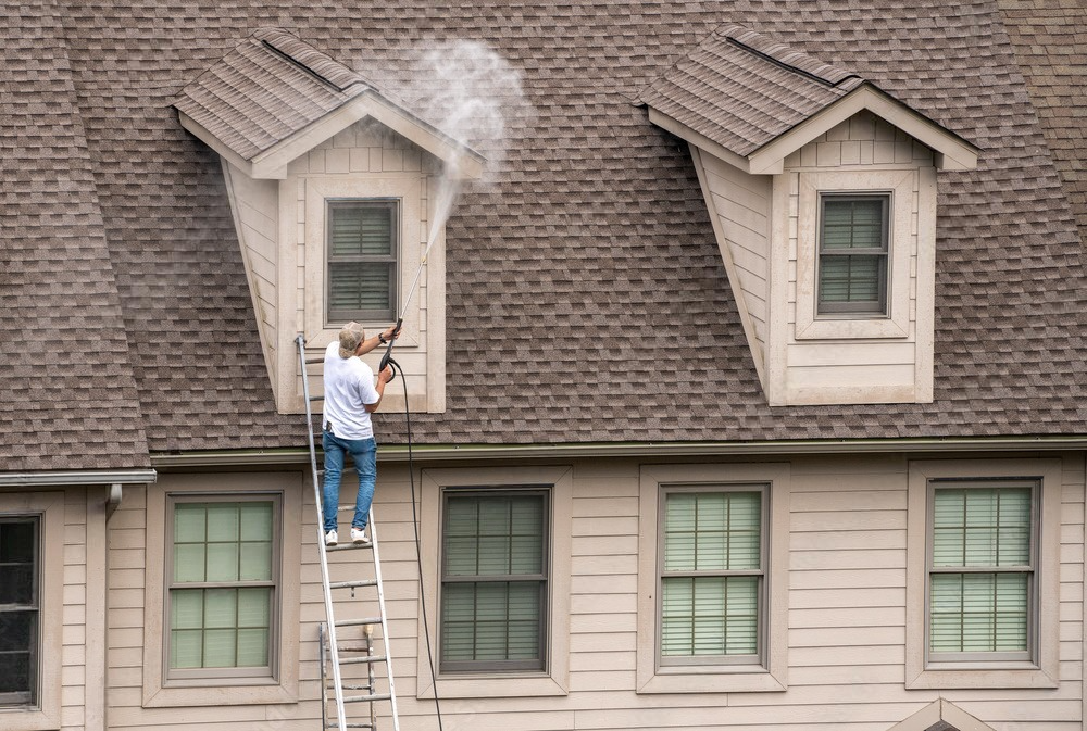 A man is standing on a ladder cleaning the side window of a house.