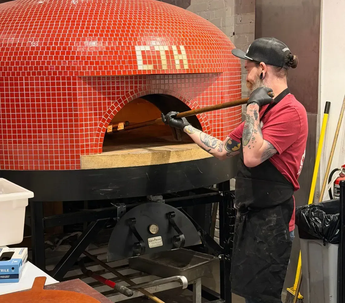 A chef using a peel to place pizza inside a red tiled brick oven.
