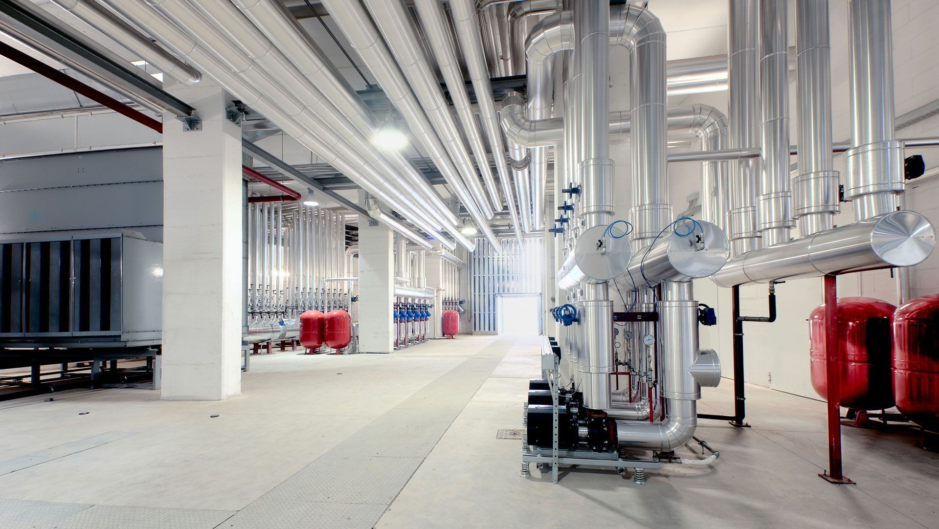 Bright industrial room with exposed pipes, ducts, and red tanks along a white corridor