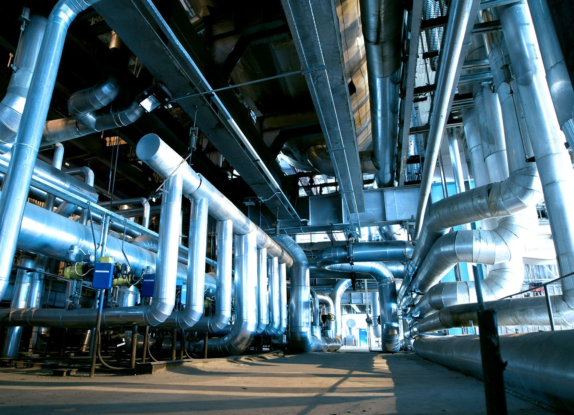 Industrial plant interior with large silver pipes and ducts overhead, lit by cool blue light.