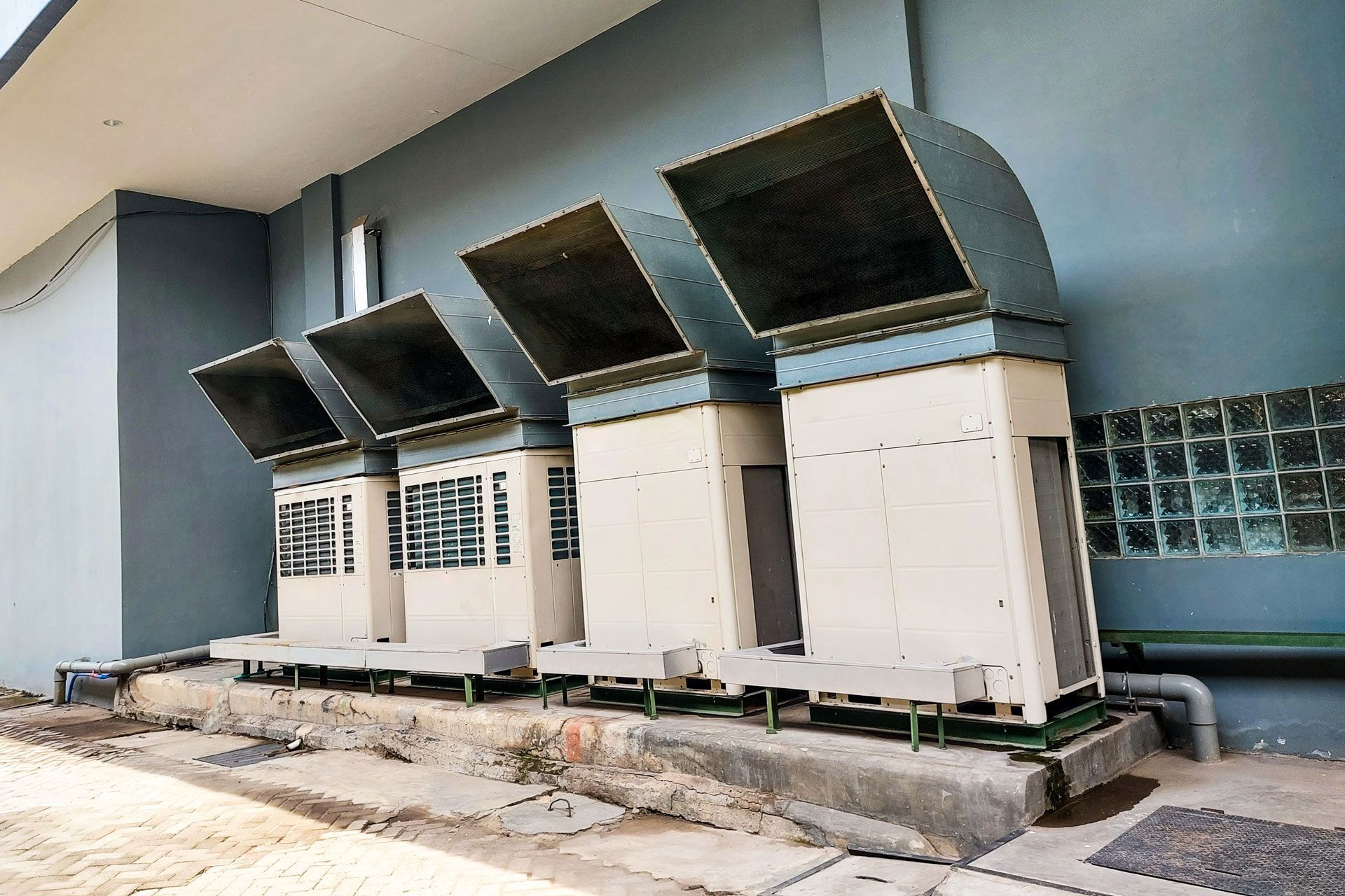 Row of outdoor air-conditioning units with large metal hoods along a building wall