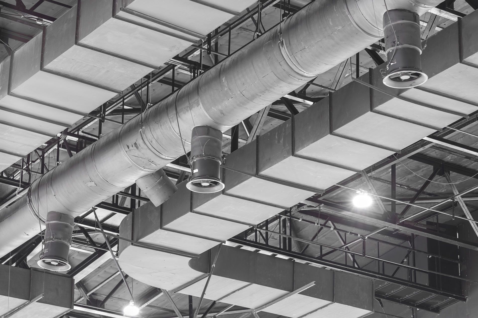 Industrial warehouse ceiling with large silver air ducts, metal trusses, and bright overhead lights