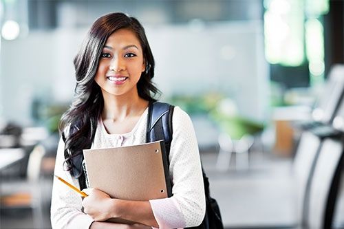 Young person smiles while holding a folder and pencil, with a backpack in a blurred indoor setting.