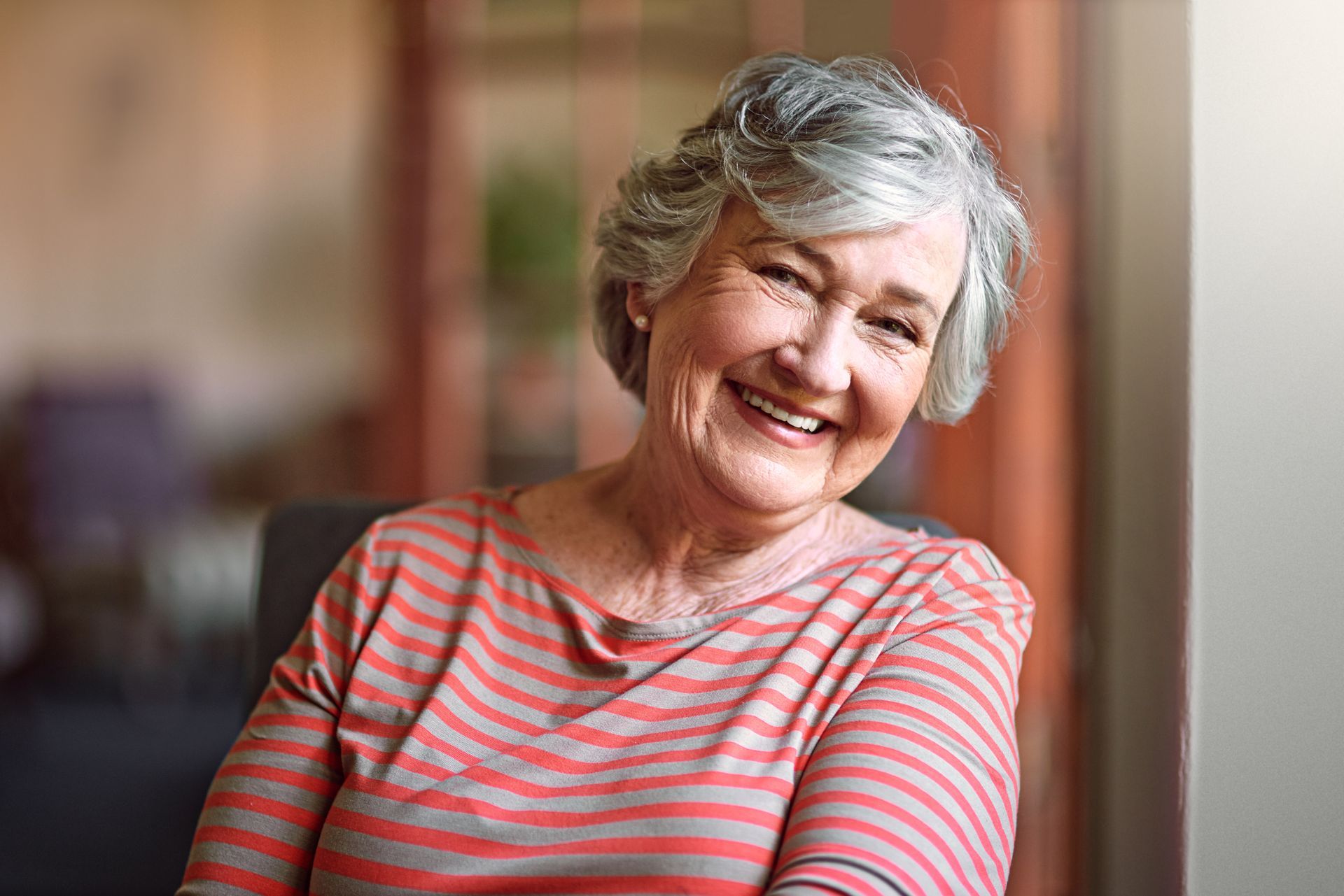 Woman with short gray hair smiles broadly, wearing a striped orange and white shirt, sitting indoors.