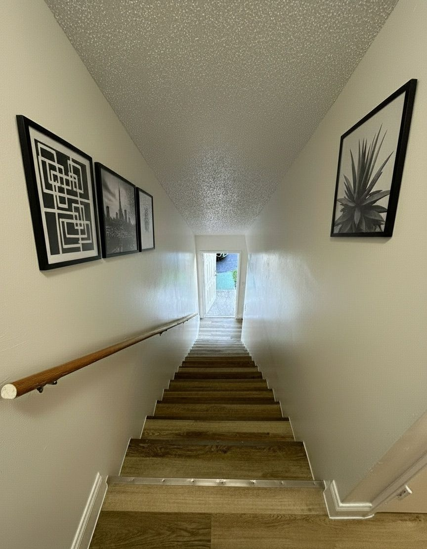 Staircase leading downwards. White walls with framed art, wood handrail, and textured ceiling. Light at the end.