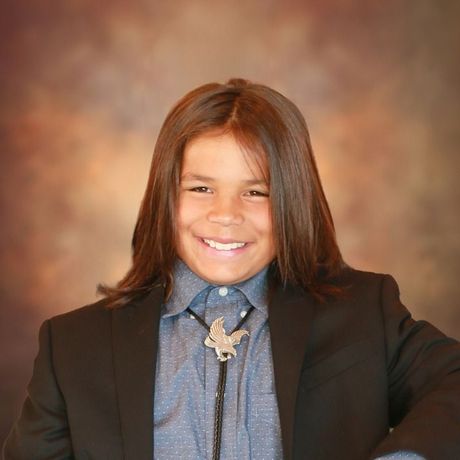 Young boy smiling, wearing a blazer, bolo tie, and button-down shirt; brown background.