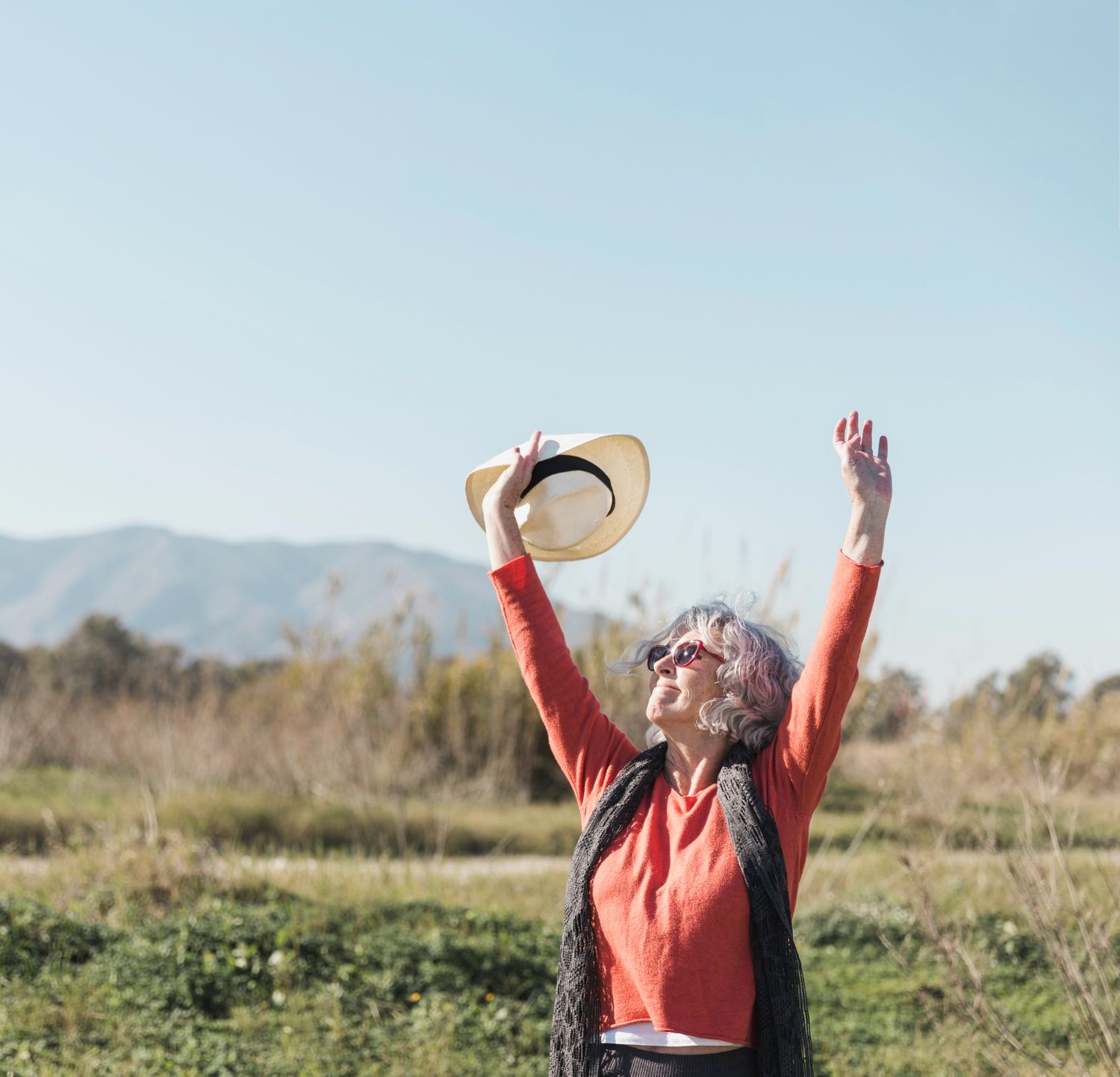 Woman in orange shirt with arms raised, holding a hat, in a field, mountains in background.