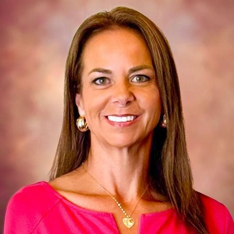 Woman with brown hair, wearing pink top, gold jewelry, smiling in front of a blurred background.