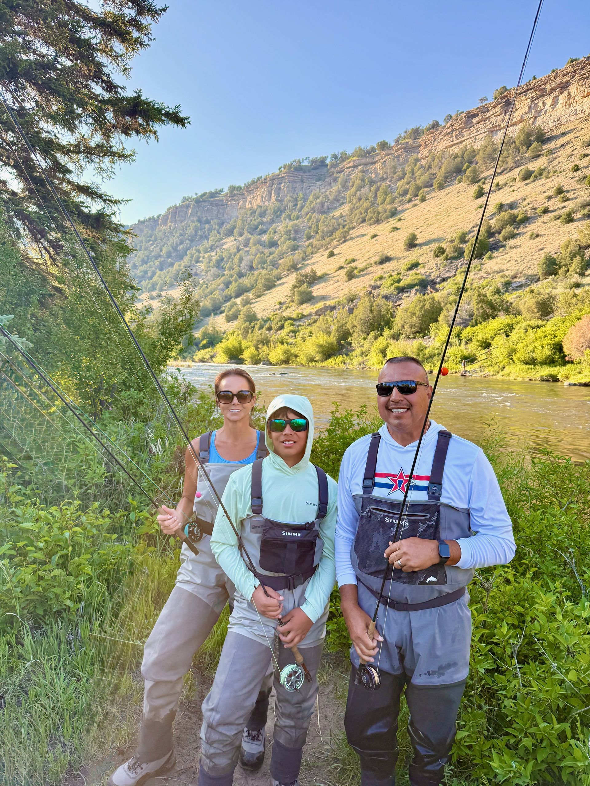 Three people fly fishing by a river, wearing waders. Mountains in the background.