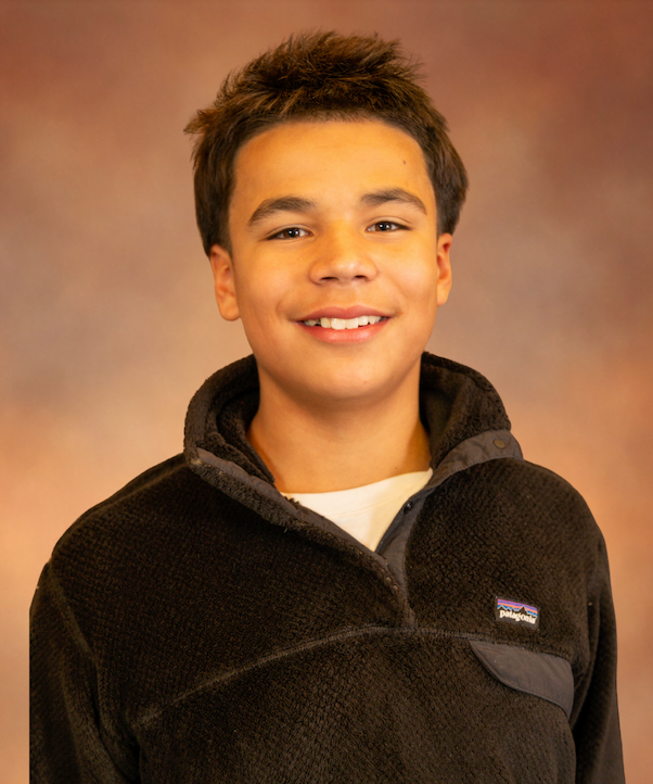 Young boy smiling, wearing a blazer, bolo tie, and button-down shirt; brown background.
