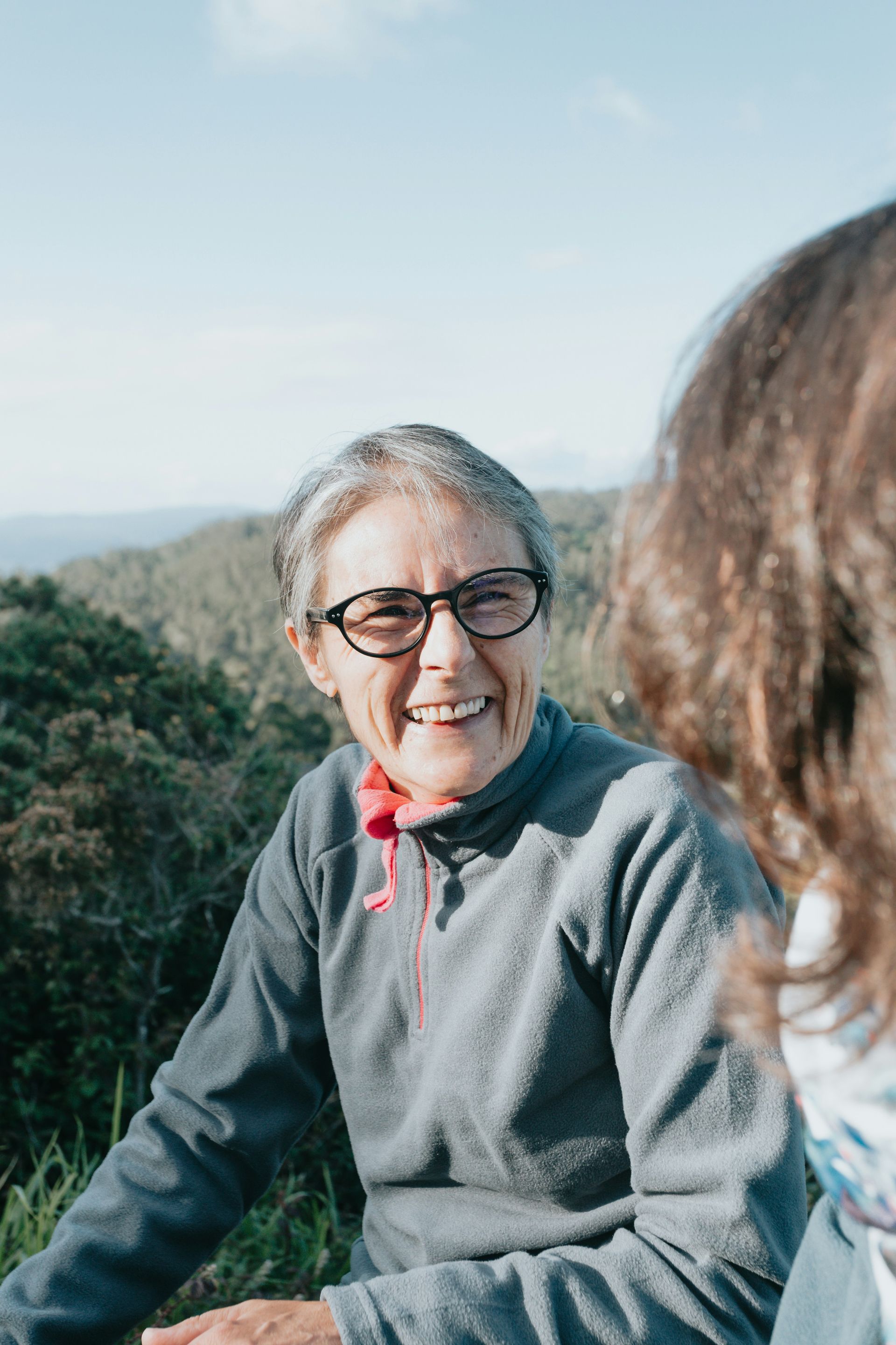 A smiling person wearing glasses and a gray fleece jacket outdoors, looking toward someone off-camera.