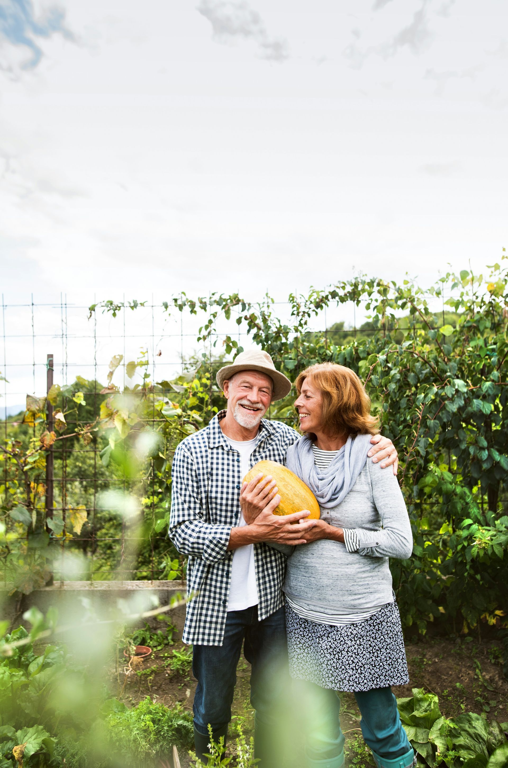 A smiling couple stands in a lush garden, holding a large yellow squash together while embracing.