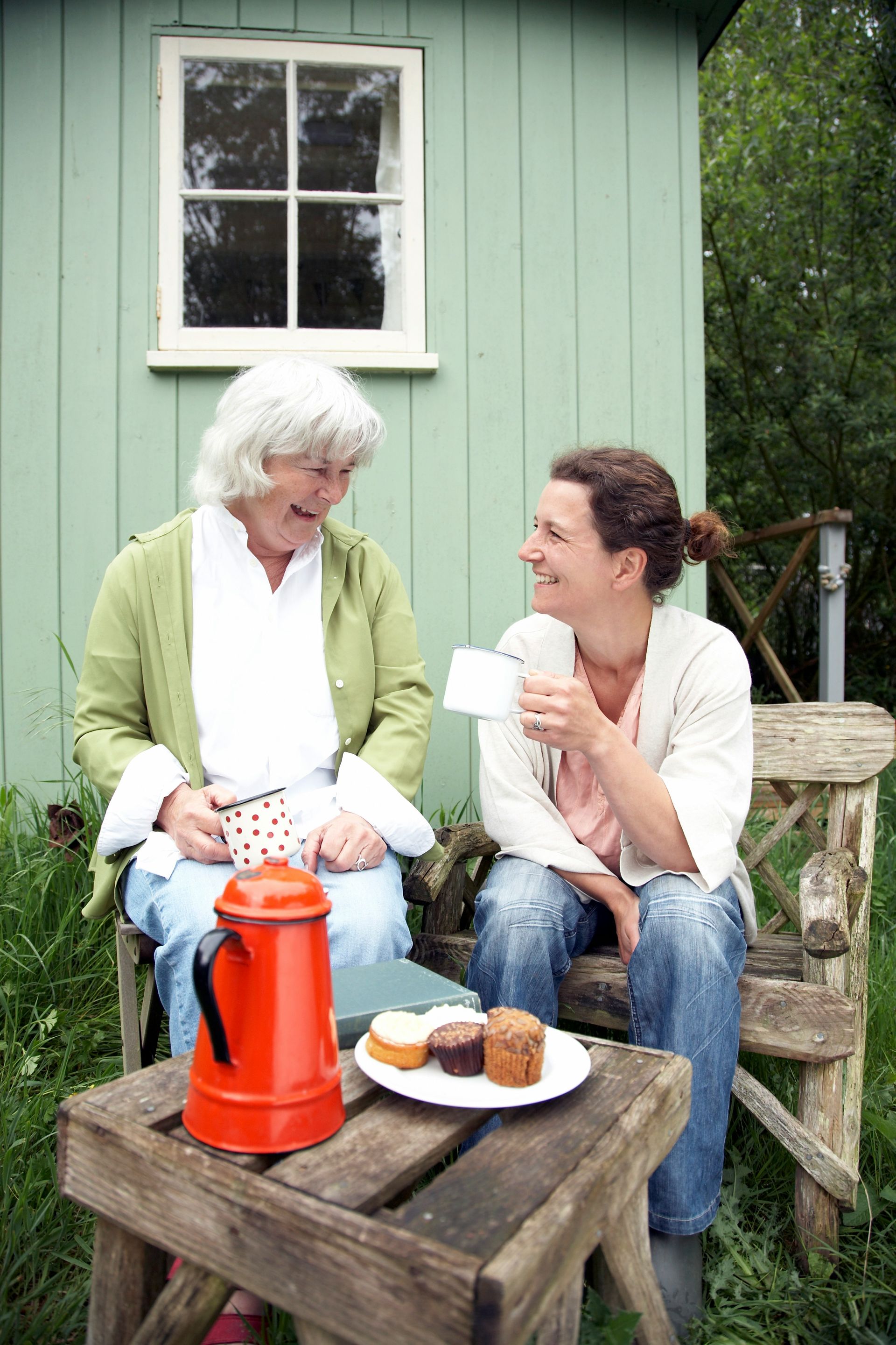 Two people sit on a wooden bench outdoors, talking and drinking tea near a red coffee pot and snacks on a small table.