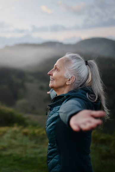 Woman in orange shirt with arms raised, holding a hat, in a field, mountains in background.