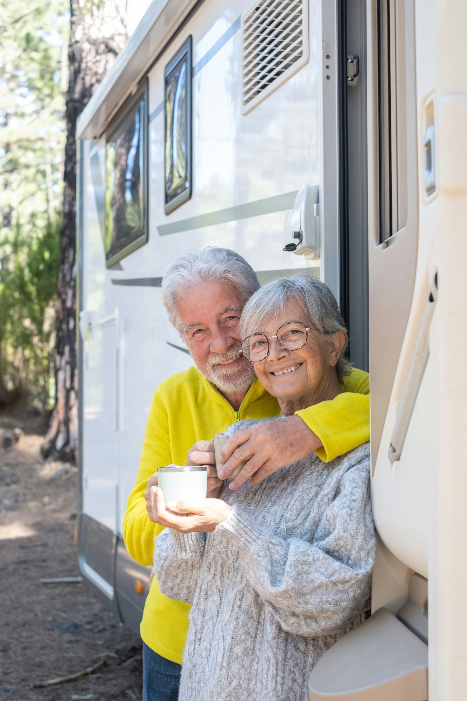 A smiling couple holding mugs while embracing in front of a camper van in a wooded setting.