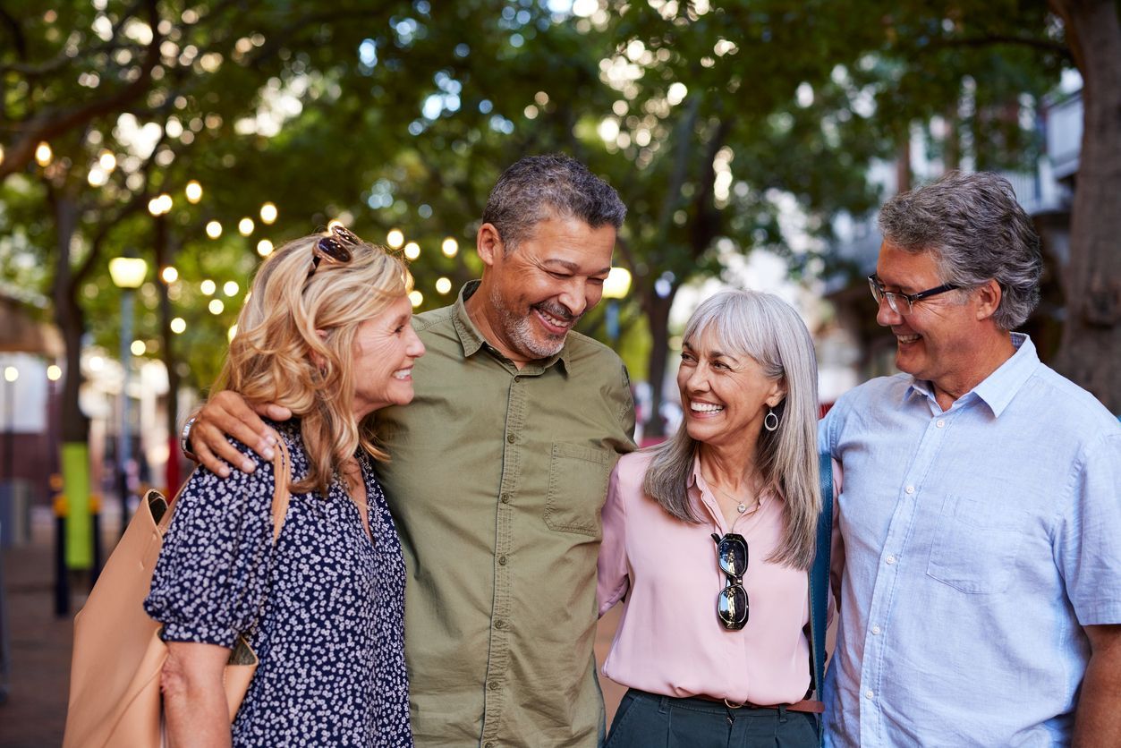 Four friends, smiling, embracing outdoors on a city street. Trees, string lights, and buildings in the background.