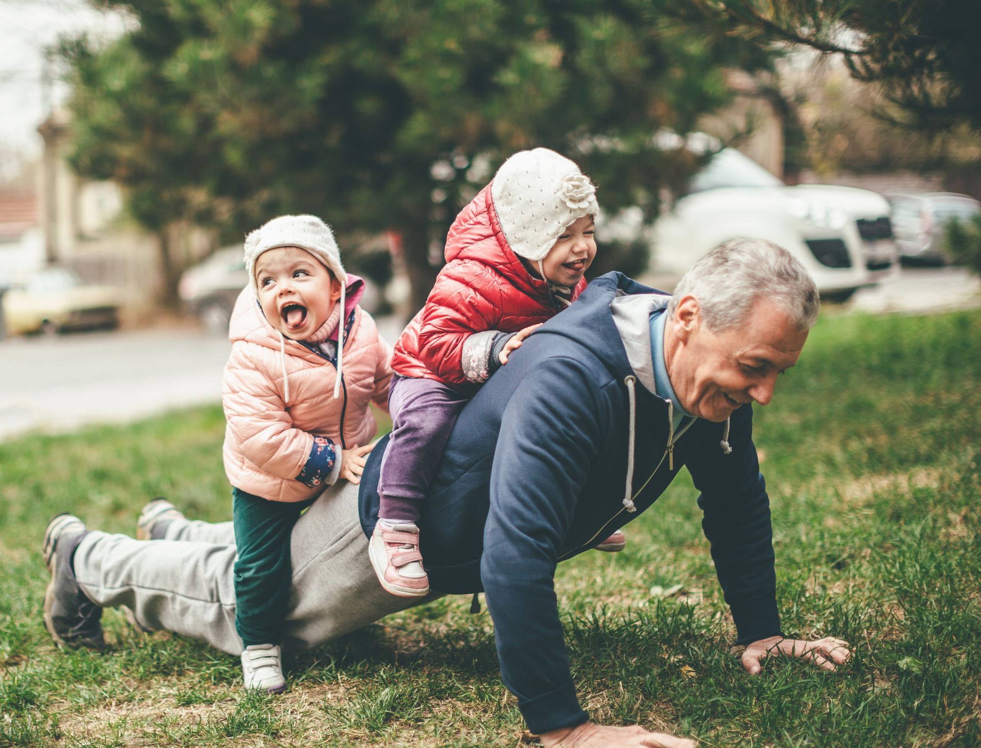 Grandfather doing push-ups with two laughing grandchildren on his back in a park.