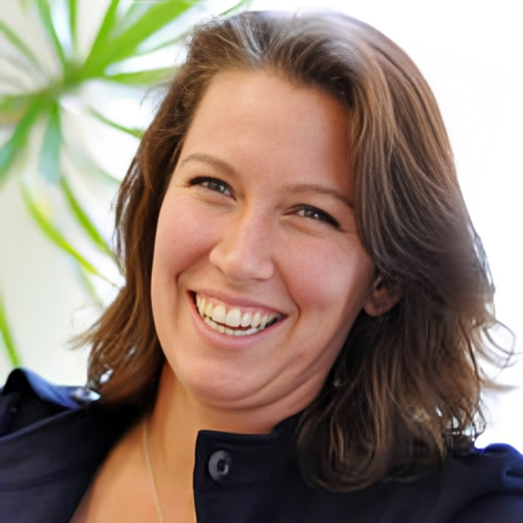 Woman with brown hair, smiling broadly, wearing a blue jacket, with a blurred background of plants.
