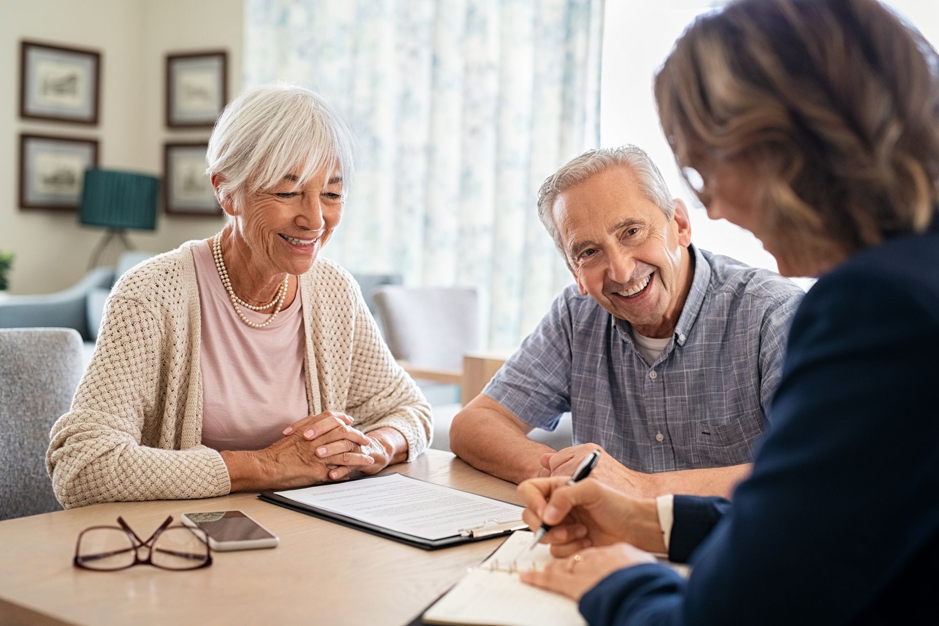 Elderly couple smiles as a person in a suit signs a document at a table in a home setting.