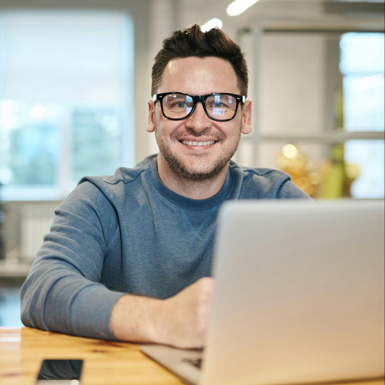 Man with glasses smiles at camera while using a laptop at a desk.