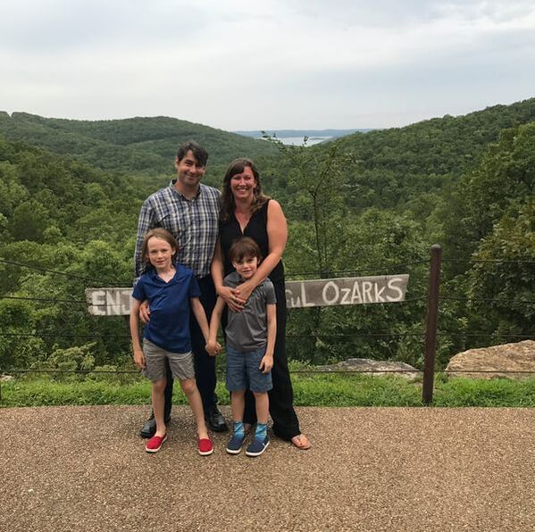 Family posing in front of a scenic Ozark Mountains view. Mountains covered in green trees, cloudy sky.