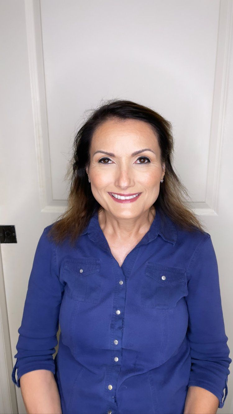 A woman in a blue shirt is smiling and standing in front of a white door.