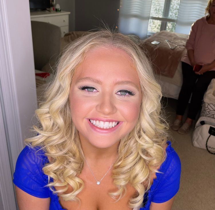 A woman with blonde curly hair is wearing a blue shirt and smiling.