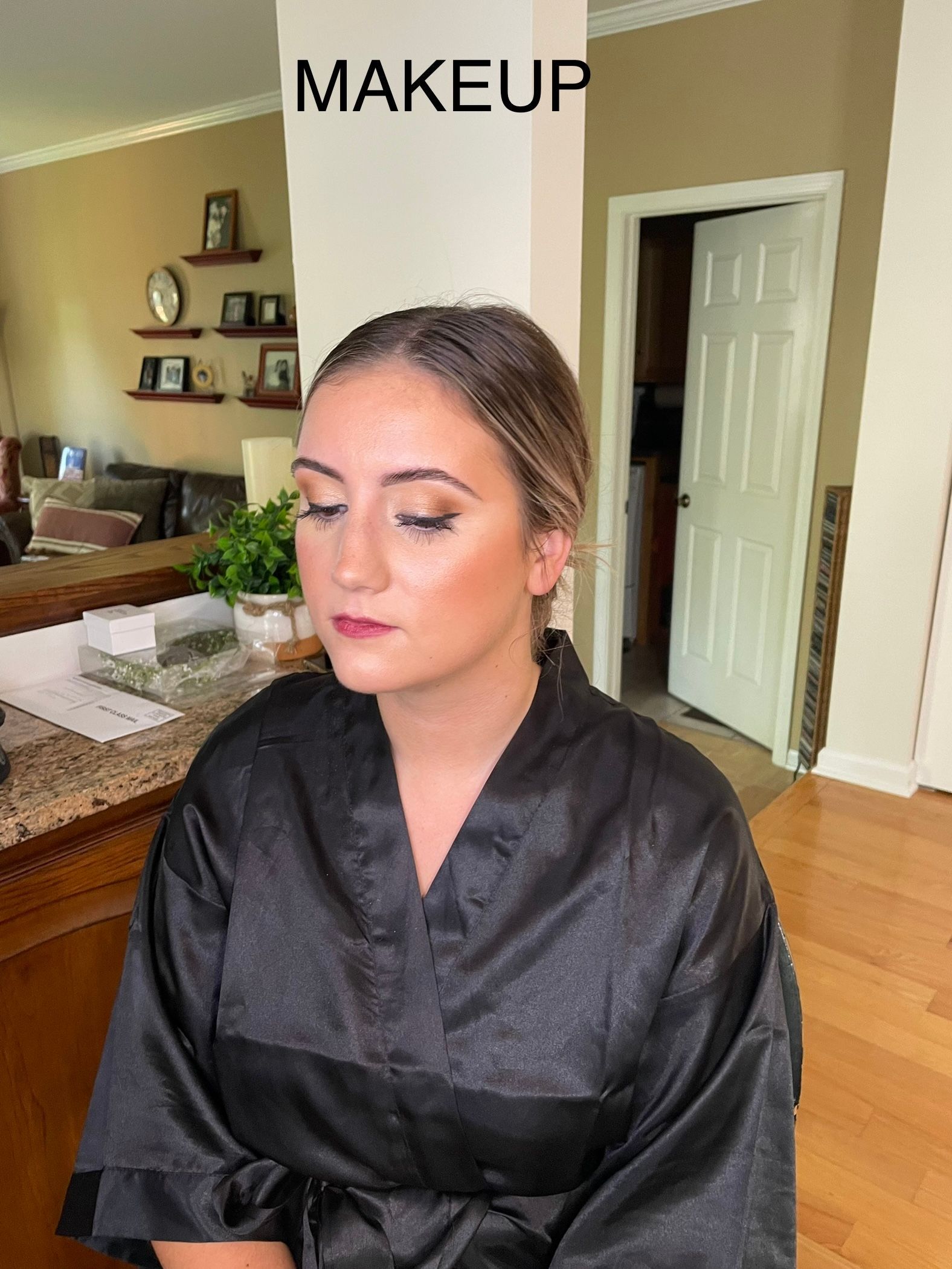 A woman in a black robe is getting her makeup done in a kitchen.