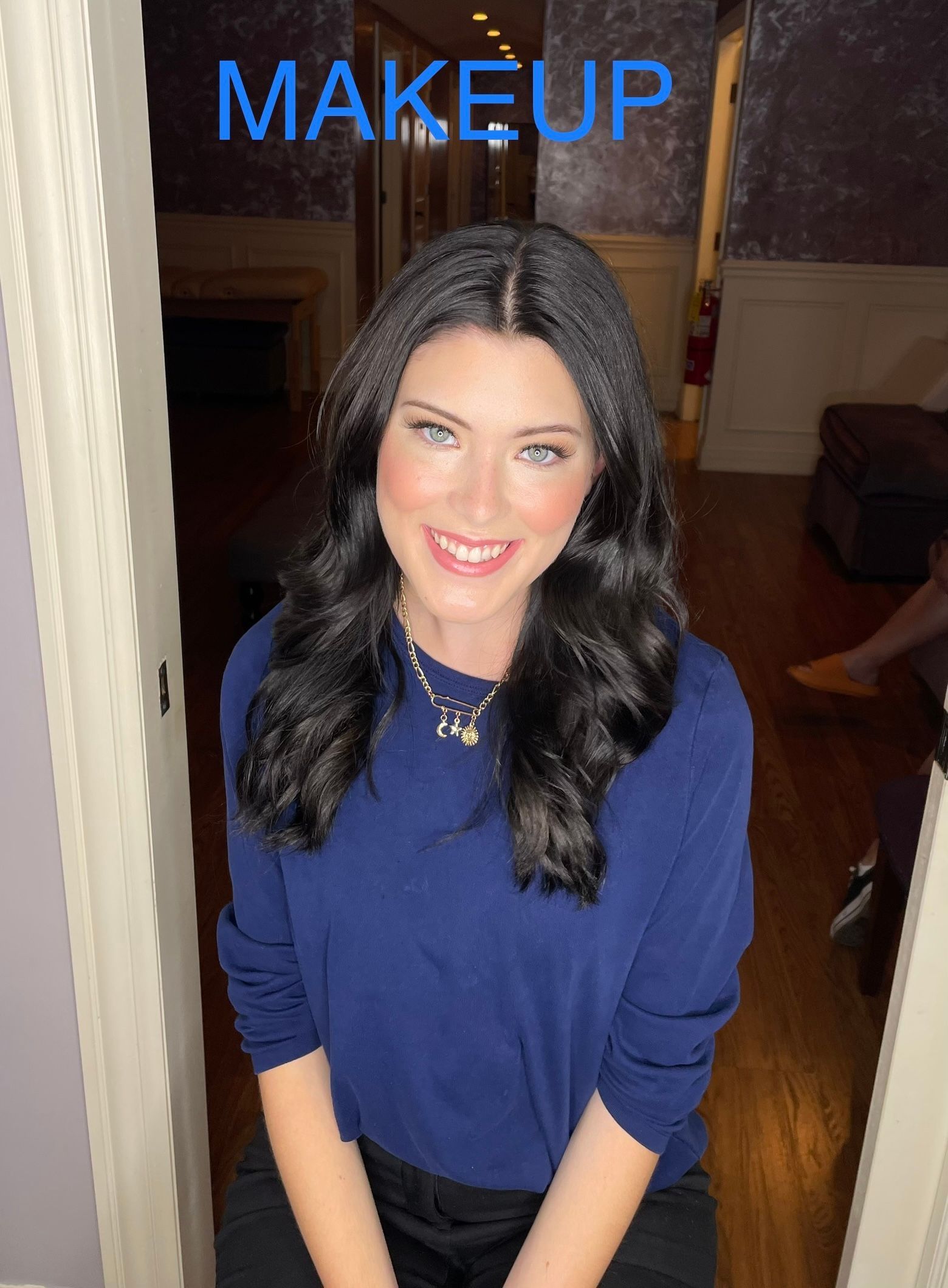 A woman in a blue shirt is smiling in front of a sign that says makeup