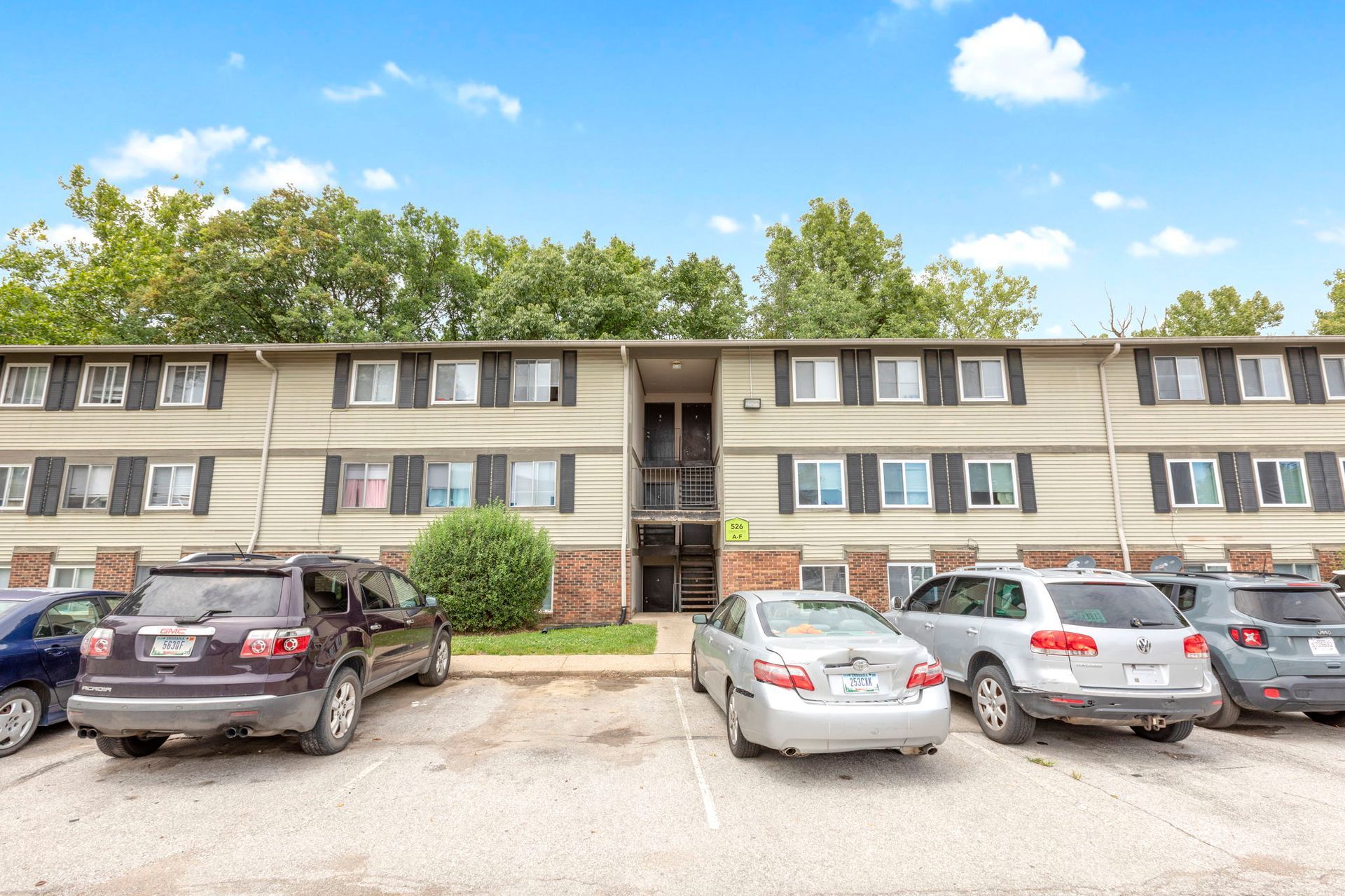Two-story apartment building with parking; cars parked out front. Light tan exterior with black shutters; sunny day.