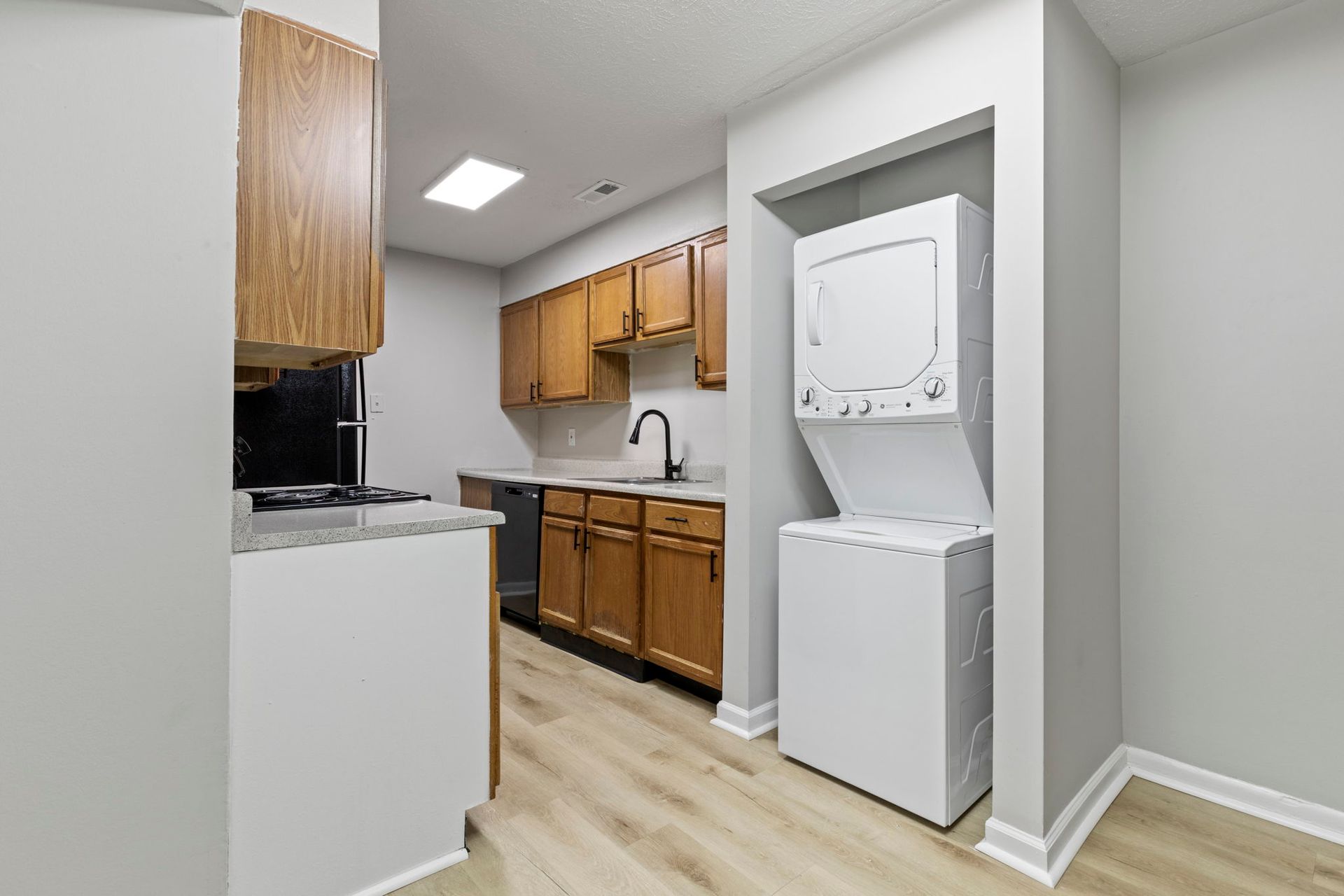 A small kitchen with wooden cabinets, light countertops, and stacked white washer/dryer units.