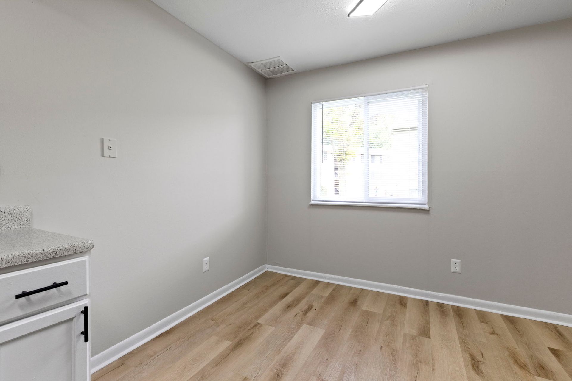 Empty room with light wood flooring, a window, and gray walls. A white cabinet with black hardware is visible.