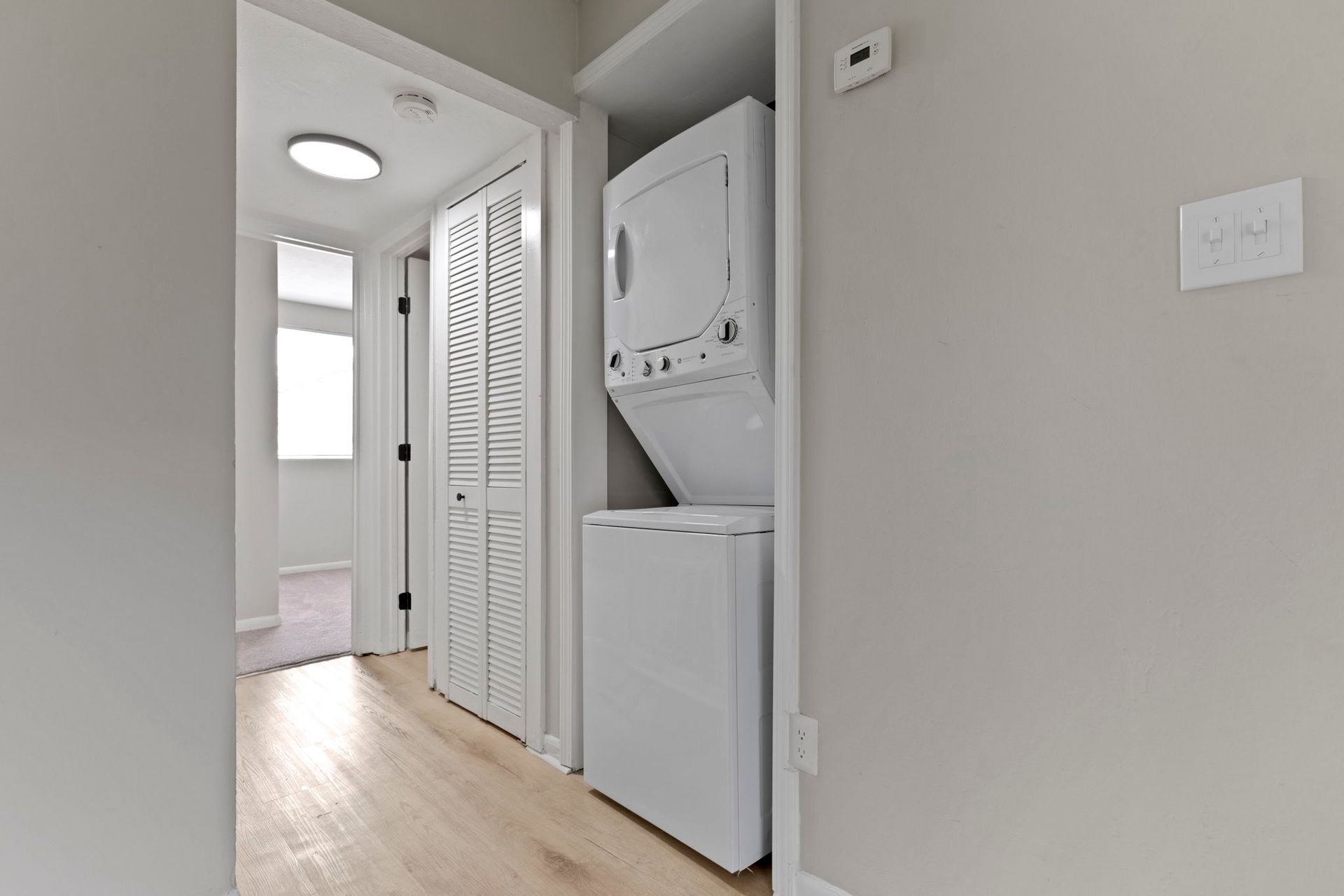 Laundry area with stacked white washer and dryer, closet with louvered doors, and hallway.