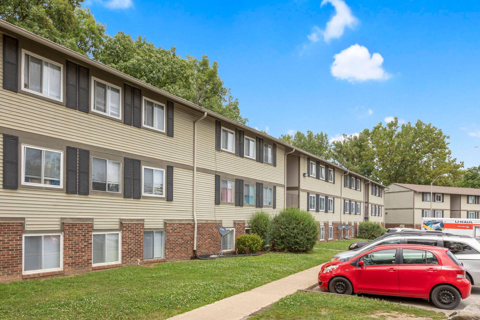 Apartment building exterior with red car parked on the right, blue sky, and green grass.