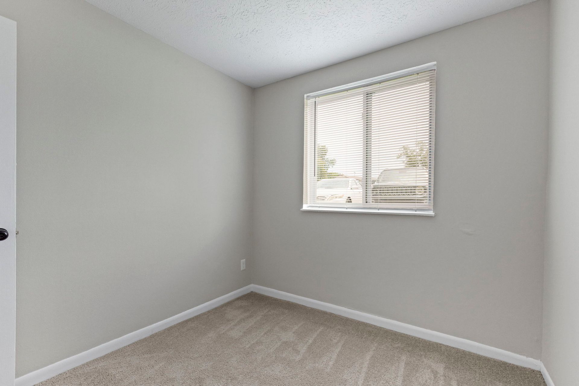 Empty bedroom with light gray walls, beige carpet, and a window with white curtains.